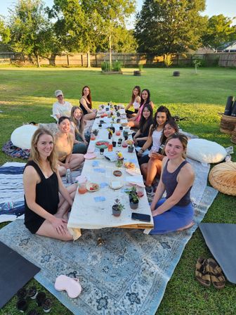 Group of friends seated on rugs and cushions around a low table for a casual outdoor backyard picnic, enjoying drinks, snacks, and small potted succulents in warm golden-hour light with trees and a wooden fence in the background.