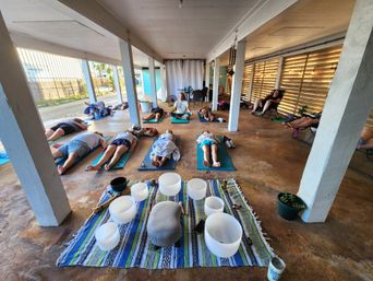 Open-air covered patio sound bath with participants reclining on yoga mats around crystal singing bowls on a colorful blanket, instructor seated in the center and a warm wooden slat wall with string lights.
