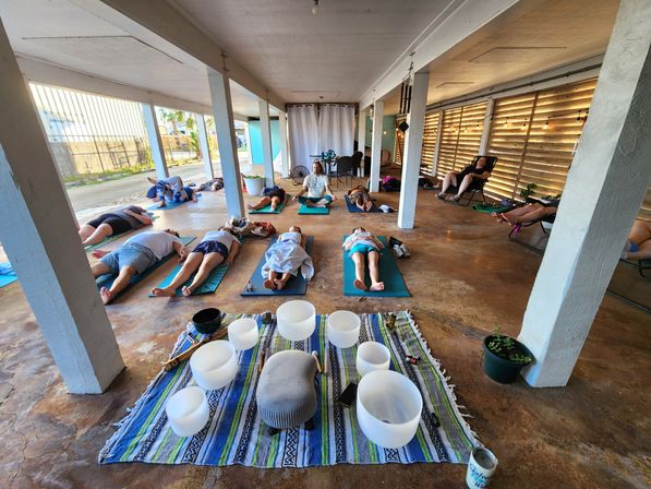 Open-air covered patio sound bath with participants reclining on yoga mats around crystal singing bowls on a colorful blanket, instructor seated in the center and a warm wooden slat wall with string lights.