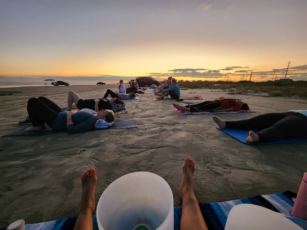 Group beach yoga and meditation on mats along a sandy shore at sunset with the ocean horizon and warm orange sky.