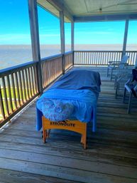 Relaxing waterfront massage table draped in blue linens on a covered wooden deck porch overlooking calm coastal waters and a bright blue sky