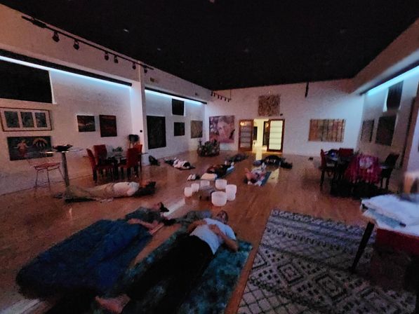 Dim, art-filled studio with people lying on mats for a sound bath or relaxation session; crystal singing bowls arranged in the center, framed paintings on white walls and soft blue accent lighting.