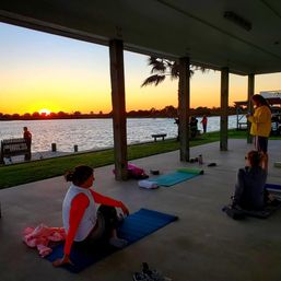 Sunset waterfront yoga on a covered pavilion — people on mats stretching near palm trees, a wooden dock and calm water under an orange sky.