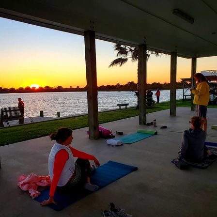 Sunset waterfront yoga on a covered pavilion — people on mats stretching near palm trees, a wooden dock and calm water under an orange sky.