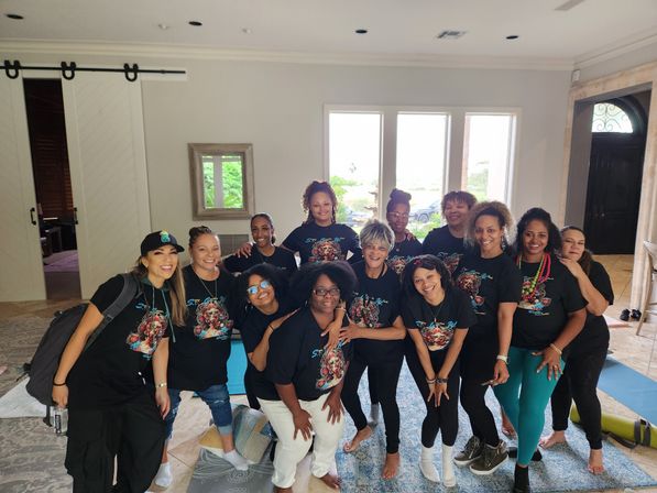 Smiling diverse group of women in matching graphic t-shirts posing in a sunlit living room with yoga mats, wellness retreat vibe