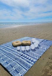 Blue-and-white patterned beach blanket on a sandy shore with two woven straw cushions and frosted crystal singing bowls arranged for a seaside meditation sound bath, calm ocean and pale blue sky on the horizon.