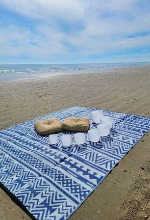 Blue-and-white patterned beach blanket on a sandy shore with two woven straw cushions and frosted crystal singing bowls arranged for a seaside meditation sound bath, calm ocean and pale blue sky on the horizon.