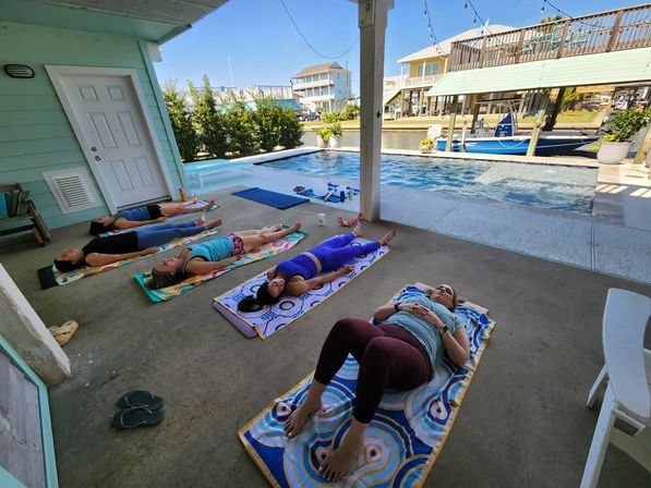 Group doing poolside yoga in savasana on colorful towels under a covered patio, overlooking a pool, boat dock and canal-side coastal houses