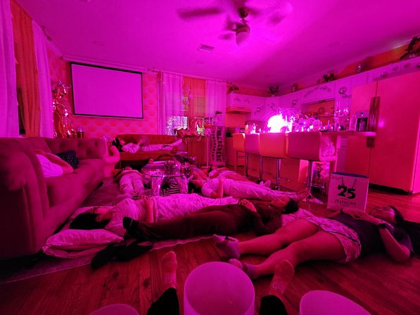 Magenta-lit living room and kitchen with a projector screen, couch and bar stools; a group of people lying on pillows across the hardwood floor in a cozy pink-lit hangout.