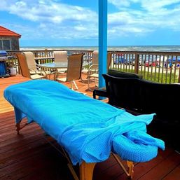 Relaxing oceanfront wooden deck with a blue-covered massage table, patio dining chairs and grill, overlooking sandy beach umbrellas, rolling waves and a bright blue sky.