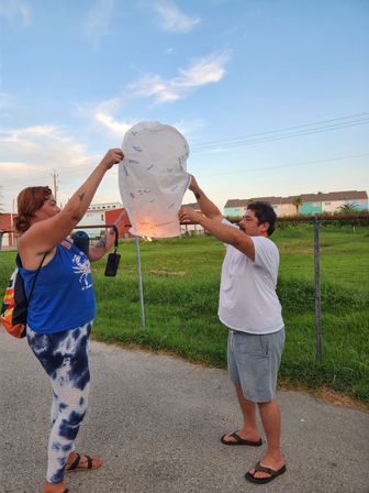 Two people releasing a glowing paper sky lantern covered in handwritten messages at dusk in a grassy residential field — woman in a blue tank and tie‑dye leggings and a man in a white t‑shirt hold the lantern as a small flame warms inside.