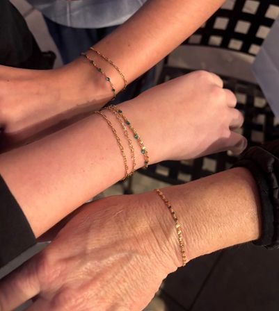 Close-up of three wrists wearing delicate gold chain bracelets with tiny blue beads, layered and touching over a black lattice chair — minimalist stacked wrist jewelry