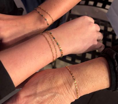 Close-up of three wrists wearing delicate gold chain bracelets with tiny blue beads, layered and touching over a black lattice chair — minimalist stacked wrist jewelry