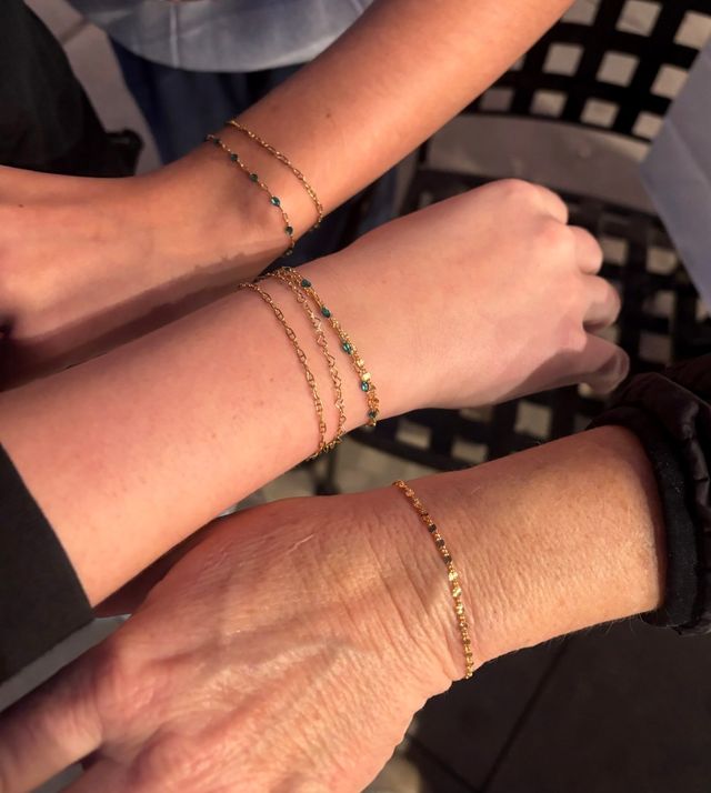 Close-up of three wrists wearing delicate gold chain bracelets with tiny blue beads, layered and touching over a black lattice chair — minimalist stacked wrist jewelry