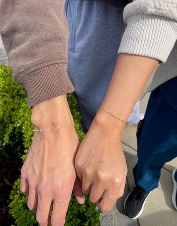 Close-up outdoor shot of two wrists side-by-side wearing delicate chain bracelets — one silver and one gold — with casual sweatshirt sleeves, green shrubs and sidewalk in the background.
