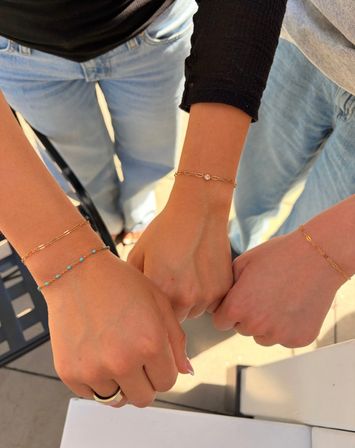 Three friends fist-bumping outdoors, close-up of wrists wearing delicate gold chain bracelets — one with tiny turquoise beads — light-wash jeans and sunlit patio in the background.