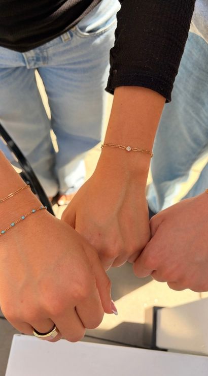 Three friends fist-bumping outdoors, close-up of wrists wearing delicate gold chain bracelets — one with tiny turquoise beads — light-wash jeans and sunlit patio in the background.