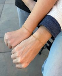 Two wrists wearing matching delicate gold chain bracelets with a denim-sleeve cuff and jeans posed over outdoor patio tiles — minimalist jewelry shot.