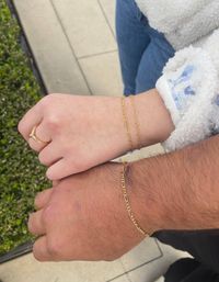 Close-up of couple holding hands outdoors, both wearing delicate matching gold chain bracelets; one hand has a dainty ring and a white fleece sleeve, tiled sidewalk and green hedge in background.