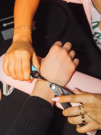 Close-up of two hands using small pliers to repair a delicate gold bracelet on a wrist, pink armrest and manicured nails suggesting a salon or jewelry repair setting