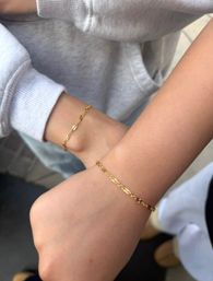 Close-up of two wrists wearing matching delicate gold chain bracelets with tiny sunburst links; one wrist in a grey sweatshirt sleeve, minimalist jewelry shot in a casual outdoor setting