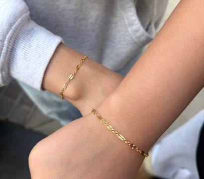 Close-up of two wrists wearing matching delicate gold chain bracelets with tiny sunburst links; one wrist in a grey sweatshirt sleeve, minimalist jewelry shot in a casual outdoor setting