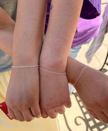 Close-up of three people linking wrists outdoors, each wearing a delicate silver chain bracelet connecting their hands, sunlit skin and a peek of a red card in one hand.