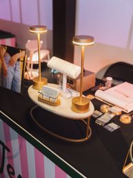 Glowing gold tabletop lamps spotlight delicate chain bracelets on a white T-bar atop a marble display tray on a pink-striped vendor table — chic jewelry display at a pop-up market