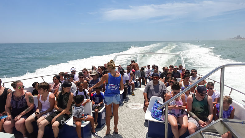 Crowded sightseeing boat tour on a sunny summer day — passengers seated on deck, crew walking between rows, white wake trailing toward the distant coastline under a clear blue sky.