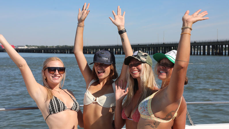 Four smiling women in bikinis and baseball caps cheering with arms raised on a sunny boat ride near a coastal bridge over calm blue water