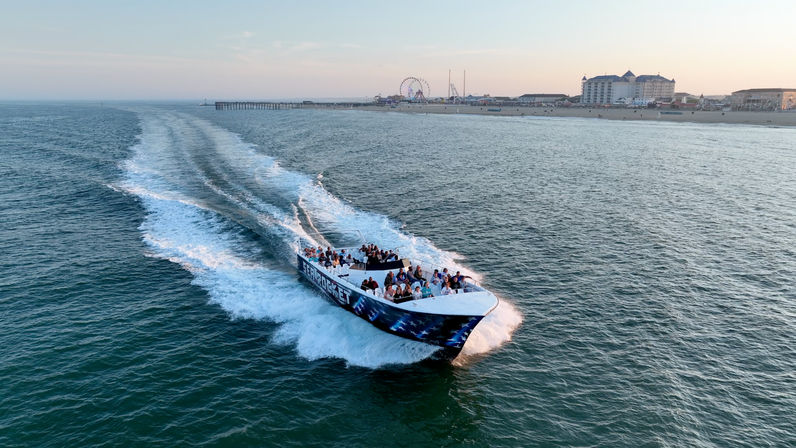 Speedboat full of passengers cutting through blue ocean, leaving a foamy wake toward a sandy beach with a long pier, Ferris wheel and seaside hotels at sunset.