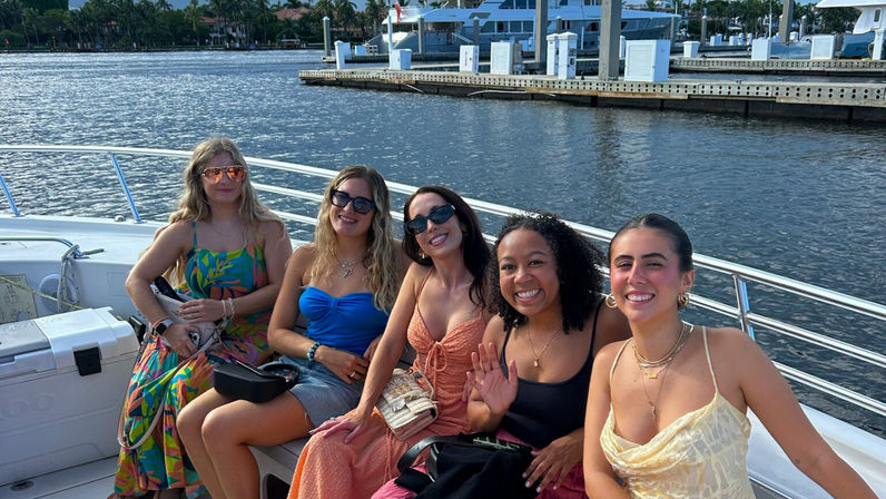 Cheerful group of five friends smiling on a white motorboat at a sunny waterfront marina, wearing summer dresses and sunglasses with docked yachts and palm-lined shore in the background.