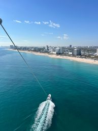 Aerial view on a sunny day of turquoise ocean and sandy beachfront with a coastal city skyline; a speedboat leaves a white wake while pulling a long towline toward the shore.