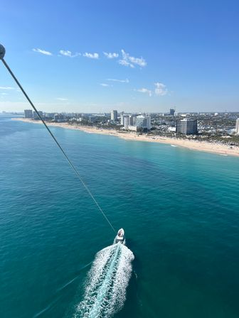 Aerial view on a sunny day of turquoise ocean and sandy beachfront with a coastal city skyline; a speedboat leaves a white wake while pulling a long towline toward the shore.