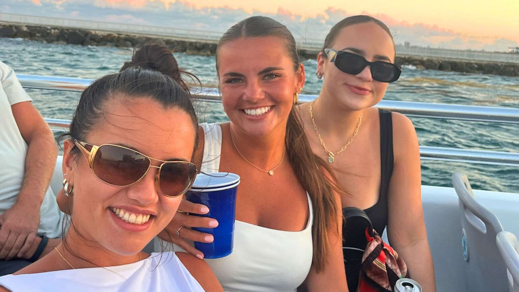 Three smiling friends on a coastal sunset boat ride, wearing sunglasses and summer outfits, one holding a blue cup and another a can.