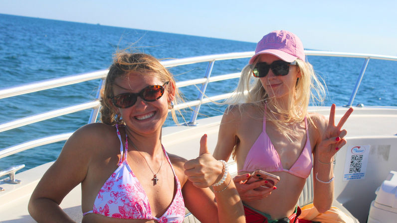 Two young women in pink bikinis and sunglasses smiling on a motorboat in open blue ocean, one giving a thumbs-up and the other flashing a peace sign on a sunny summer boating trip.