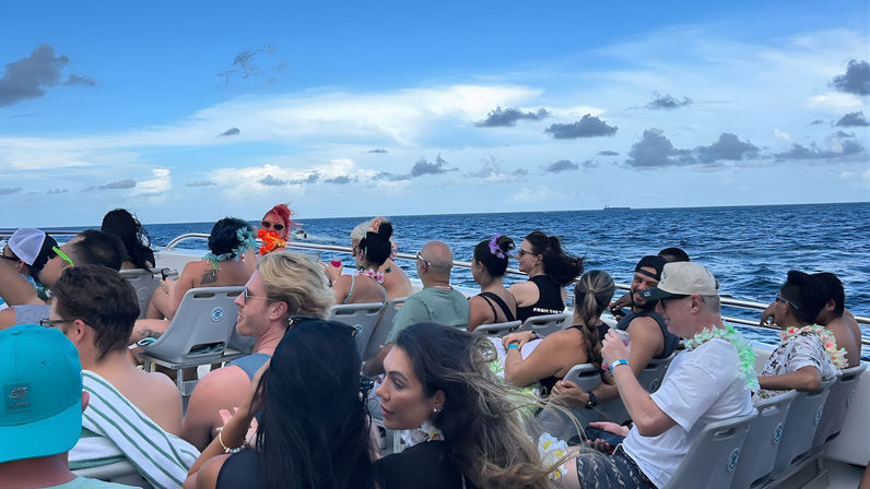 Group of people on a daytime party boat cruise over a deep blue ocean, wearing leis and sunglasses and enjoying the sea breeze under a bright sky with scattered clouds