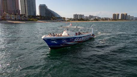 Patriotic blue-and-red tour boat with American flag and passengers cruising offshore near a beachfront high-rise skyline on a sunny coastal day.