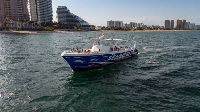 Patriotic blue-and-red tour boat with American flag and passengers cruising offshore near a beachfront high-rise skyline on a sunny coastal day.