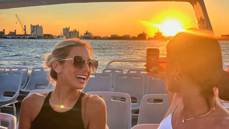 Two friends on a harbor sunset boat cruise — one in sunglasses laughing while the other snaps a smartphone photo, ships and cranes on the waterlined skyline.