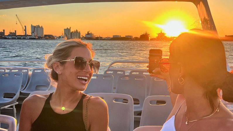 Two friends on a harbor sunset boat cruise — one in sunglasses laughing while the other snaps a smartphone photo, ships and cranes on the waterlined skyline.