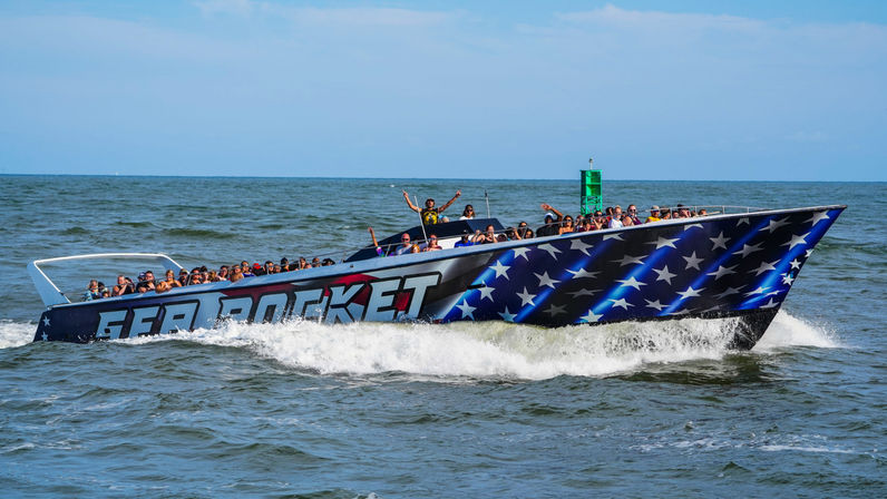 Star-spangled speedboat full of cheering passengers on a sunny ocean tour, speeding through coastal waters and leaving a white foamy wake.