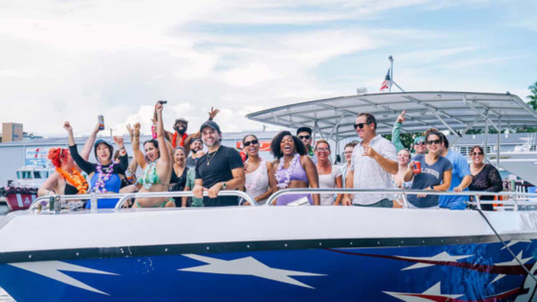 Energetic group of people cheering on a blue, star-painted party boat at a sunny marina, wearing swimsuits, leis and sunglasses and holding drinks