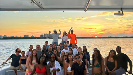 Large diverse group enjoying a sunset cruise on a boat, posing in front of calm bay waters with a distant coastal skyline and golden-orange sky.