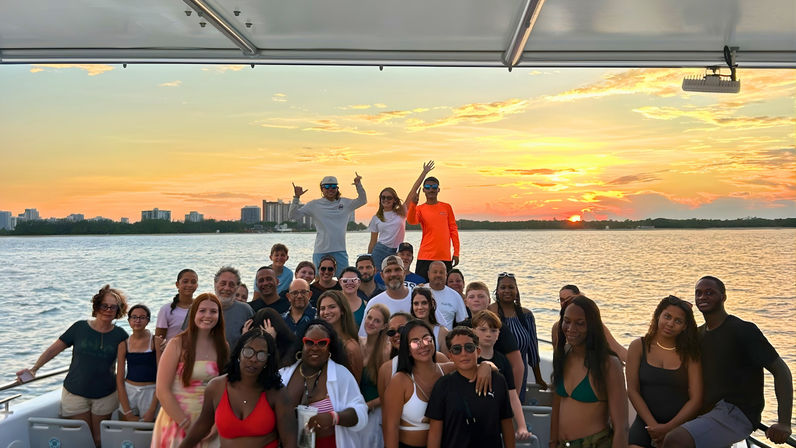 Large diverse group enjoying a sunset cruise on a boat, posing in front of calm bay waters with a distant coastal skyline and golden-orange sky.