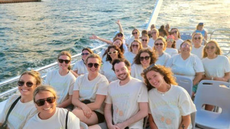 Cheery group of adults in matching light t-shirts and sunglasses seated on a boat deck during a sunny coastal cruise, smiling with ocean water and sunset glow in the background.