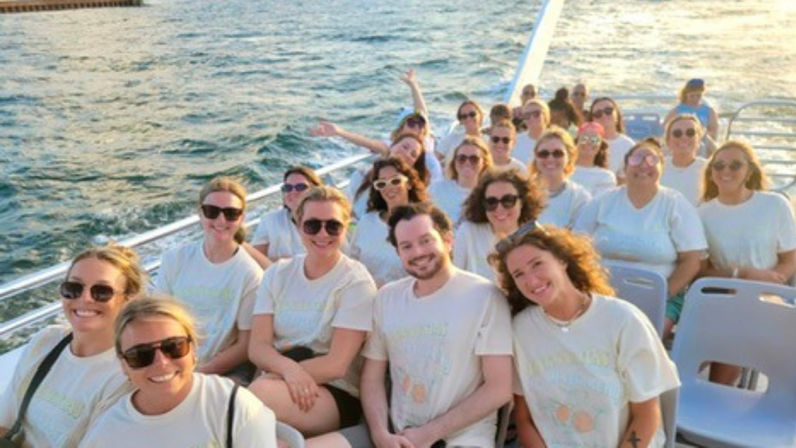 Cheery group of adults in matching light t-shirts and sunglasses seated on a boat deck during a sunny coastal cruise, smiling with ocean water and sunset glow in the background.