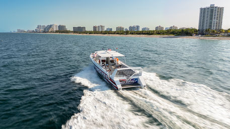 Powerboat speeding across deep-blue ocean near a palm-lined beachfront and high-rise condos, kicking up a dramatic white wake under clear sunny skies.
