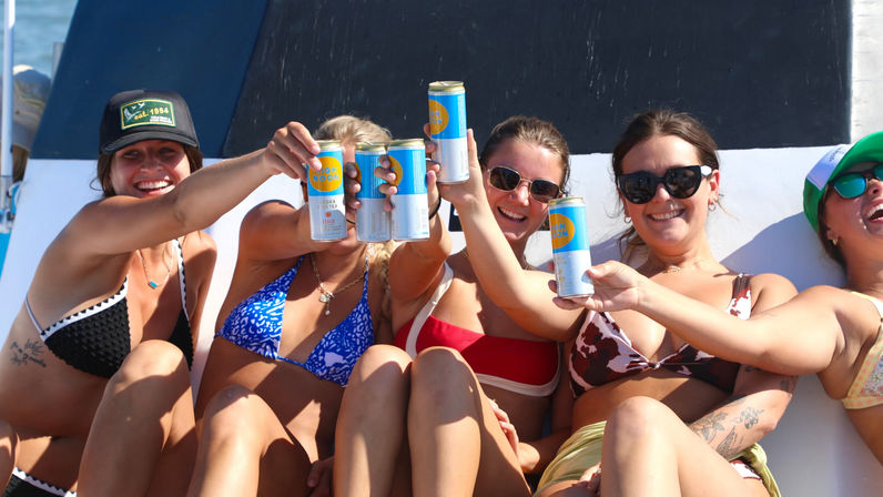 Group of friends in bikinis on a sunny boat, smiling and toasting with canned cocktails over the ocean for a summer beach party vibe