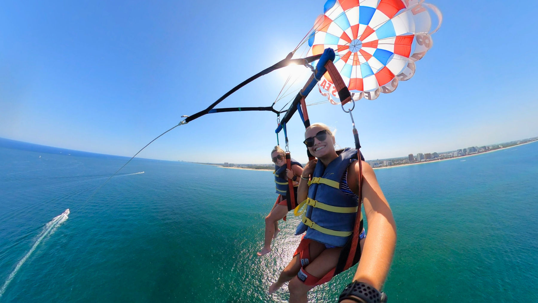Two smiling parasail riders high above turquoise ocean, tethered to a red, white and blue parachute with a sandy coastline and beachfront skyline below on a sunny day